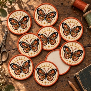 May include: A collection of circular embroidered patches featuring a butterfly design with orange, black, and white wings, accented by small stars and sunburst rays on a cream-colored fabric background with a stitched orange border.