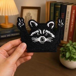 May include: A black and white embroidered patch featuring a raccoon with its arms raised in a peace sign gesture. The design shows the raccoon's face with detailed white stitching for the eyes, ears, and whiskers against a solid black background.