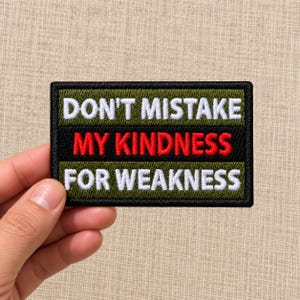 May include: A rectangular embroidered patch with a black border, featuring an olive green background and the text DON'T MISTAKE in white, MY KINDNESS in red, and FOR WEAKNESS in white, all stitched in a bold, capitalized sans-serif font.