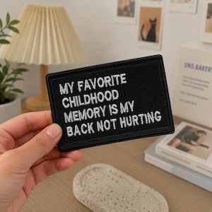 May include: A rectangular black embroidered patch featuring white text that reads: MY FAVORITE CHILDHOOD MEMORY IS MY BACK NOT HURTING. The patch has a clean, stitched border and a flat, fabric-based construction with clear, legible lettering.