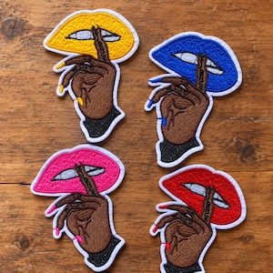 May include: Four embroidered patches featuring a brown hand with long colored nails held up to lips. The lips are yellow, blue, pink, and red. Each patch has a white border and depicts a hand making a shushing gesture against vibrant lips.