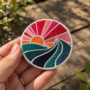May include: A circular embroidered patch featuring a stylized landscape design. The upper portion displays a bright orange sun with radiating beams in shades of pink and red. The lower portion depicts rolling hills in teal, dark blue, and green.
