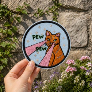 May include: Embroidered patch featuring an orange cat with green eyes, emitting pink laser beams from its eyes. The text "PEW PEW" is embroidered in green. The patch has a light blue background with embroidered stars and a black border.