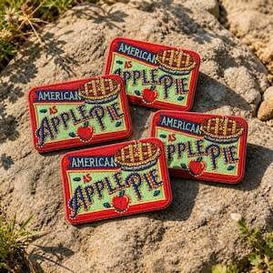 May include: Four embroidered patches with the text "AMERICAN AS APPLE PIE". Each patch features a red border, a green background, and a pie and apple illustration. The text is in blue and gold.