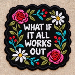 May include: A black embroidered patch with a scalloped edge featuring the white text WHAT IF IT ALL WORKS OUT in the center. The text is surrounded by a colorful floral wreath with pink roses, white daisies, green leaves, and small red berries.