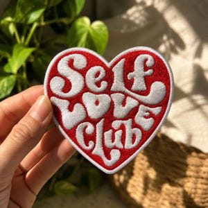 May include: A heart-shaped embroidered patch with a red background and white stitched lettering that reads Self Love Club in a bold, stylized font. The patch features a thick white border and a textured surface created by the embroidery thread.