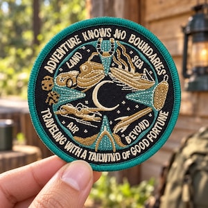 May include: A circular embroidered patch with a teal border and black background. It features gold thread illustrations of a ship, plane, and compass, with the text ADVENTURE KNOWS NO BOUNDARIES and TRAVELING WITH A TAILWIND OF GOOD FORTUNE.
