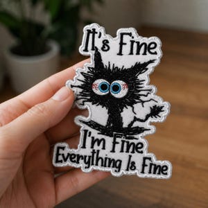 May include: An embroidered patch featuring a chaotic, black, spiky-furred cat with wide, bloodshot blue eyes. The white background of the patch displays the black embroidered text: It's Fine, I'm Fine, Everything Is Fine, arranged above and below.