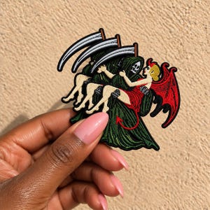 May include: An embroidered patch featuring a grim reaper in a dark green hooded robe holding a silver scythe, carrying a blonde figure with red wings and a red dress. The design includes a small red devil tail and black border stitching.