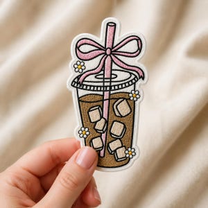 May include: An embroidered patch featuring an iced coffee in a clear plastic cup with a pink straw and a pink bow tied around it. The design includes several white daisies with yellow centers and light-colored ice cubes inside the brown liquid.