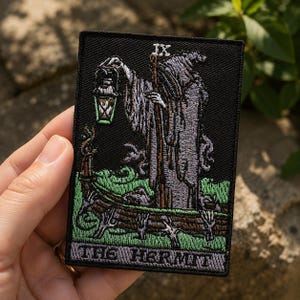 May include: A rectangular embroidered patch featuring a tarot card design of The Hermit. The figure wears a hooded cloak, holds a staff and a glowing lantern, and stands on a log above green grass. The Roman numeral IX and text THE HERMIT appear.