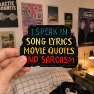 May include: A black embroidered patch with a scalloped edge featuring the text I SPEAK IN in teal, SONG LYRICS MOVIE QUOTES in yellow, and AND SARCASM in red. The patch has a dense stitched texture and a rectangular shape with rounded corners.