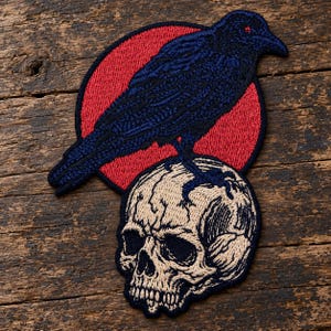 May include: An embroidered patch featuring a dark blue raven with a small red eye perched atop a beige human skull. The raven is positioned in front of a solid red circular background. The entire design is outlined in a thick black border.