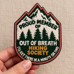 May include: An embroidered patch shaped like a mountain peak with a dark green background, white snow-capped mountains, and pine trees. It features the text PROUD MEMBER OF OUT OF BREATH HIKING SOCIETY and I'LL GET THERE IN A MINUTE OR TWO.