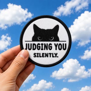 May include: A circular embroidered patch with a white background and a black border. The design features a black cat silhouette peeking over a horizontal line, with the black text "JUDGING YOU SILENTLY." printed below the cat's eyes.