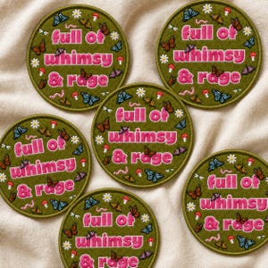 May include: Five circular olive green embroidered patches featuring the pink text full of whimsy & rage. Each patch is decorated with small white daisies, red mushrooms, pink worms, and various colorful butterflies scattered across the surface.