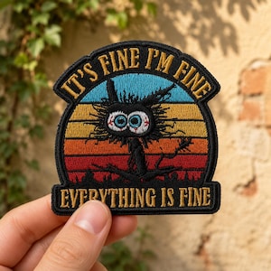 May include: An embroidered patch featuring a black, disheveled cat with wide, bloodshot eyes centered against a sunset background of blue, yellow, orange, and red stripes. The text reads IT'S FINE I'M FINE at the top and EVERYTHING IS FINE below.