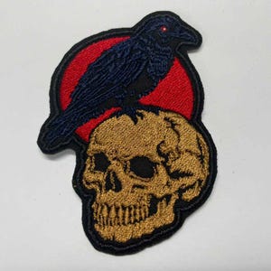 May include: An embroidered patch featuring a dark blue raven with a single red eye perched atop a tan human skull. The design is set against a circular red background, all outlined with a thick black border, creating a high-contrast graphic.
