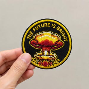 May include: A circular embroidered patch with a black background and a yellow border. The center features a colorful mushroom cloud graphic in shades of red, orange, and yellow. The text THE FUTURE IS BRIGHT is stitched in yellow along the top edge.