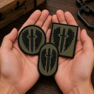 May include: Three olive green embroidered patches featuring a black winged sword design. The set includes one circular patch, one square patch, and one oval patch, all with black stitched borders and a consistent tactical aesthetic.