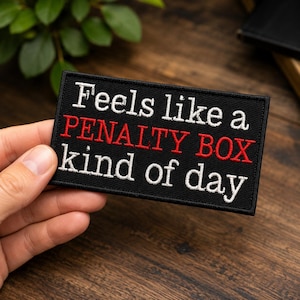 May include: A rectangular black embroidered patch with a stitched border. The text is embroidered in white and red thread, reading "Feels like a PENALTY BOX kind of day" with the words "PENALTY BOX" highlighted in bold red capital letters.