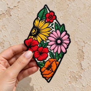 May include: A diamond-shaped embroidered patch featuring a vibrant floral arrangement. The design includes a large yellow sunflower, a pink daisy, three red hibiscus-style flowers, and an orange blossom, all accented with green leaves.