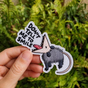 May include: An embroidered patch featuring a gray opossum with a wide-open mouth, a pink tongue, and sharp teeth. The patch includes the black text "DON'T TELL ME TO SMILE" arranged in a speech bubble shape above the opossum's head on a white background.