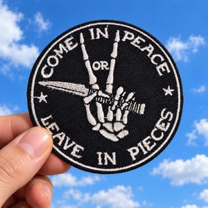 May include: A circular black embroidered patch featuring a white skeleton hand making a peace sign while holding a dagger. The text "COME IN PEACE OR LEAVE IN PIECES" is stitched in white around the border with two small stars.