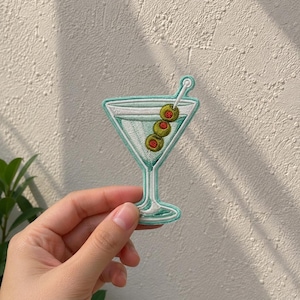 May include: An embroidered patch shaped like a martini glass with a light blue border. The glass contains a pale blue liquid and a cocktail pick holding three green olives with red centers. The design features detailed stitching throughout.