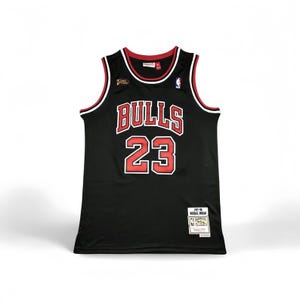 Könnte beinhalten: Schwarzes Basketballtrikot mit roten und weißen Akzenten. Der Schriftzug "BULLS" ist in weißen Blockbuchstaben mit roter Umrandung und die Nummer "23" in Rot gehalten. Ein kleines Patch mit Text ist sichtbar.