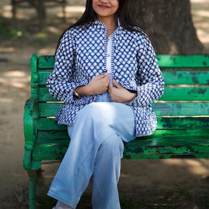 May include: A person wearing a blue and white patterned jacket, white shirt, and light blue jeans. The jacket has a geometric design. The person is sitting on a green bench. White trainers complete the outfit. The text "PINKCITY HANDCRAFTED" is visible.