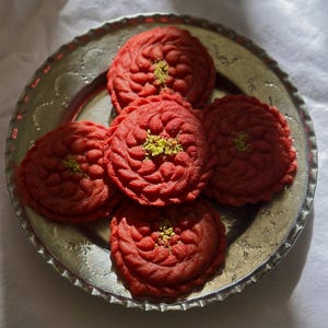 May include: A silver plate holds five red, flower-shaped cookies. Each cookie has a textured surface and is topped with a sprinkle of green pistachios. The cookies are arranged on a silver plate with an ornate edge.