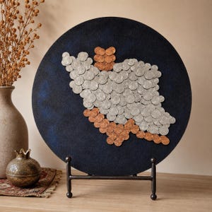 May include: A circular dark blue wall art piece featuring a map outline created from numerous silver and copper-colored coins. The artwork is displayed on a black metal stand. The coins are arranged to form a specific geographic shape.