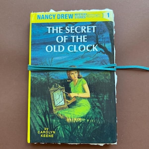 May include: A copy of the book The Secret of the Old Clock by Carolyn Keene, part of the Nancy Drew Mystery Stories series. The cover features a green-clad figure with a clock in a dark landscape, wrapped in a thin teal leather cord.