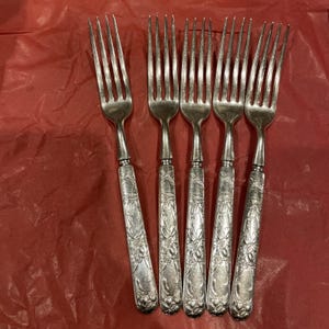 Puede incluir: Un conjunto de cinco tenedores antiguos de plata con detalles florales y de volutas ornamentales en los mangos. Los tenedores están dispuestos sobre una superficie roja texturizada, mostrando su elegante diseño y atractivo vintage.