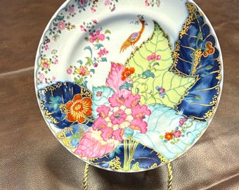 May include: A round porcelain plate featuring a vibrant floral and leaf pattern in shades of pink, yellow, blue, and orange, accented with gold trim. A small bird is depicted perched on a large leaf, with a scalloped edge and floral border.