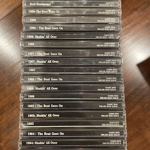 Könnte beinhalten: Ein Stapel klarer CD-Hüllen mit Classic-Rock-Alben aus den 1960er Jahren. Titel wie "Rock Renaissance", "The Beat Goes On" und "Shakin' All Over" sind zu sehen. Die CDs sind auf einer Holzoberfläche gestapelt.