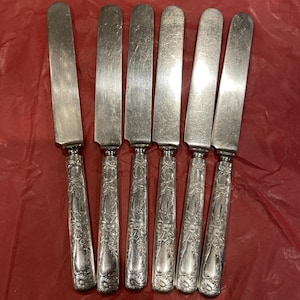 Puede incluir: Seis cuchillos de mantequilla plateados antiguos con mangos ornamentados con motivos florales y de volutas. Las hojas son rectangulares y muestran signos de desgaste. Los cuchillos están sobre una superficie roja.