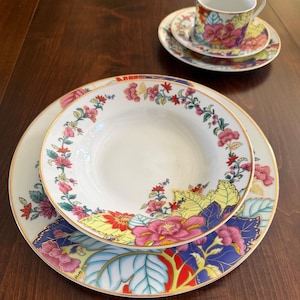 May include: A white porcelain dinnerware set featuring a vibrant floral pattern with pink, red, yellow, and blue leaves and blossoms. The set includes a large dinner plate, a smaller bowl, and a matching teacup and saucer, all with gold trim.