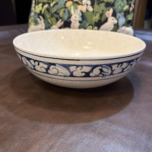May include: A round ceramic bowl with a cream-colored crackle glaze finish. The exterior features a decorative band of blue painted rabbits and scrolling foliage patterns. The bowl has a wide, open top and a smooth, curved base design.