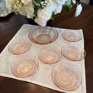 May include: A set of seven pink depression glass bowls featuring a delicate embossed floral pattern along the rims. The collection includes one larger central bowl and six smaller matching bowls, all displaying a translucent light pink hue.