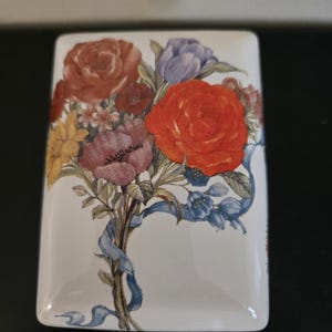 Op de afbeelding: Een rechthoekige, witte keramische doos met een bloemenboeketontwerp. Het boeket bevat rode, oranje, paarse en gele bloemen, vastgebonden met een blauw lint. De doos heeft een glanzende afwerking.