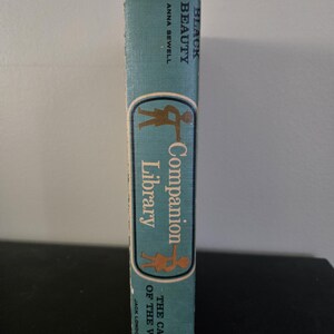 May include: A vintage book spine with the titles "Black Beauty" by Anna Sewell and "The Call of the Wild" by Jack London. The spine is teal with a gold and brown illustration and text for "Companion Library".