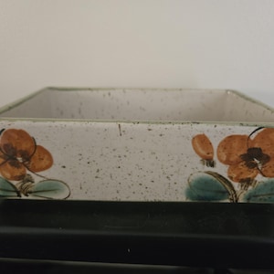 May include: Rectangular ceramic dish with a speckled off-white finish. The dish features brown and orange floral designs with green leaves on the sides. The rim has a green border. This decorative dish is suitable for serving or display.