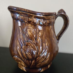 May include: A brown ceramic pitcher with a floral relief design. The pitcher has a curved spout and a handle. The glaze has a glossy finish with darker brown accents. The pitcher is likely vintage or antique.