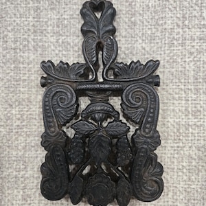 May include: Ornate black metal trivet with a handle featuring a heart-shaped cutout. The trivet showcases intricate floral and scrollwork designs, including leaves, grapes, and a central flower. The decorative piece is likely used for protecting surfaces from hot cookware.
