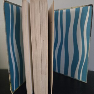 May include: An open book with pages fanned out, revealing a spine and interior pages. The covers feature a blue and white wavy stripe pattern. The book is set against a dark surface.