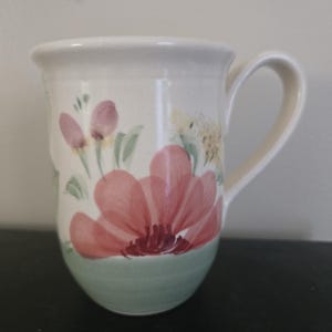 May include: A ceramic mug with a cream-colored base featuring a hand-painted floral design. The mug displays a large pink flower at the bottom, smaller pink buds, and yellow accents. The lower portion of the mug is finished in a soft sage green.