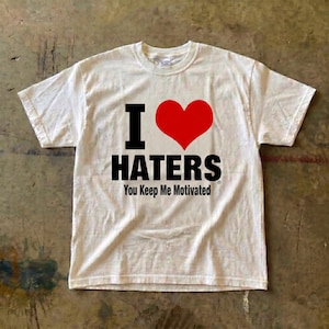 May include: White t-shirt featuring the text "I ❤️ HATERS" in black, with a red heart symbol. Underneath, the phrase "You Keep Me Motivated" is printed. The shirt is made of a soft material. The t-shirt is a casual wear item.