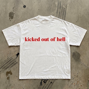 以下が含まれることがあります： 赤い文字で「kicked out of hell」と書かれた白いTシャツ。Tシャツはクルーネックで半袖です。柔らかく快適な素材で作られています。